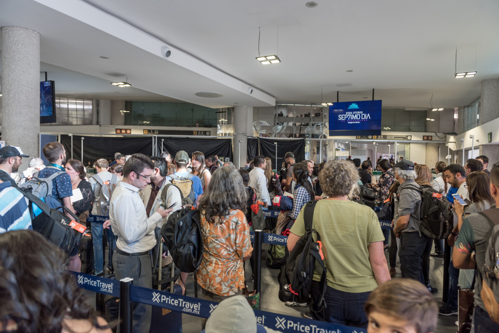 Crowded airport check-in area with people lined up, carrying luggage, and waiting at counters.