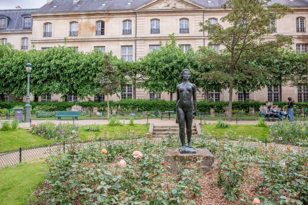 Bronze statue of a standing woman in a garden with roses and an old building in the background.