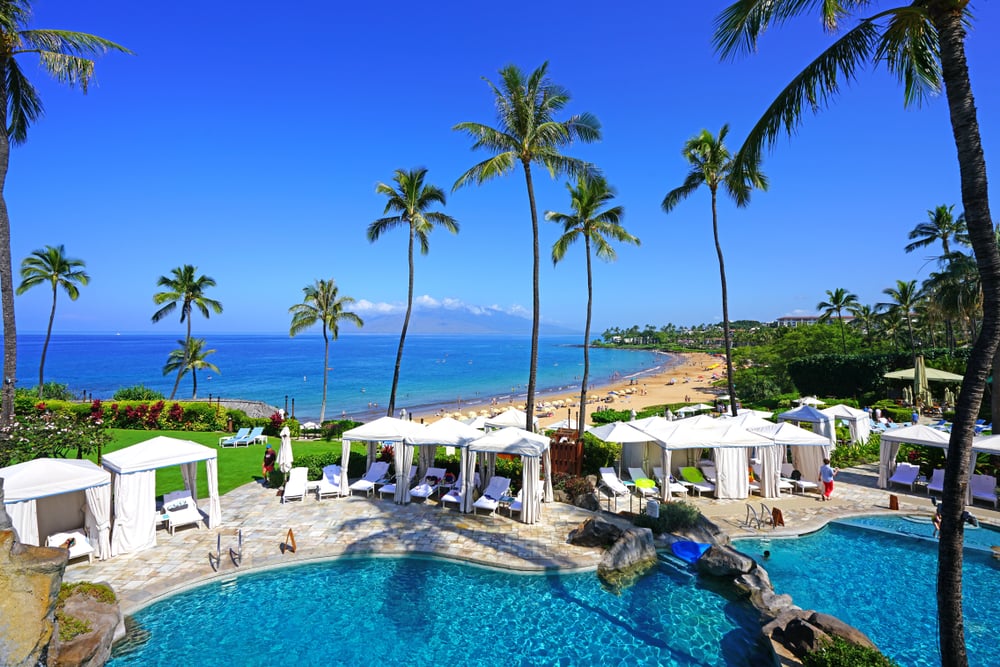 Palm trees surround a pool area with white cabanas, overlooking a sandy beach and ocean under a clear sky.