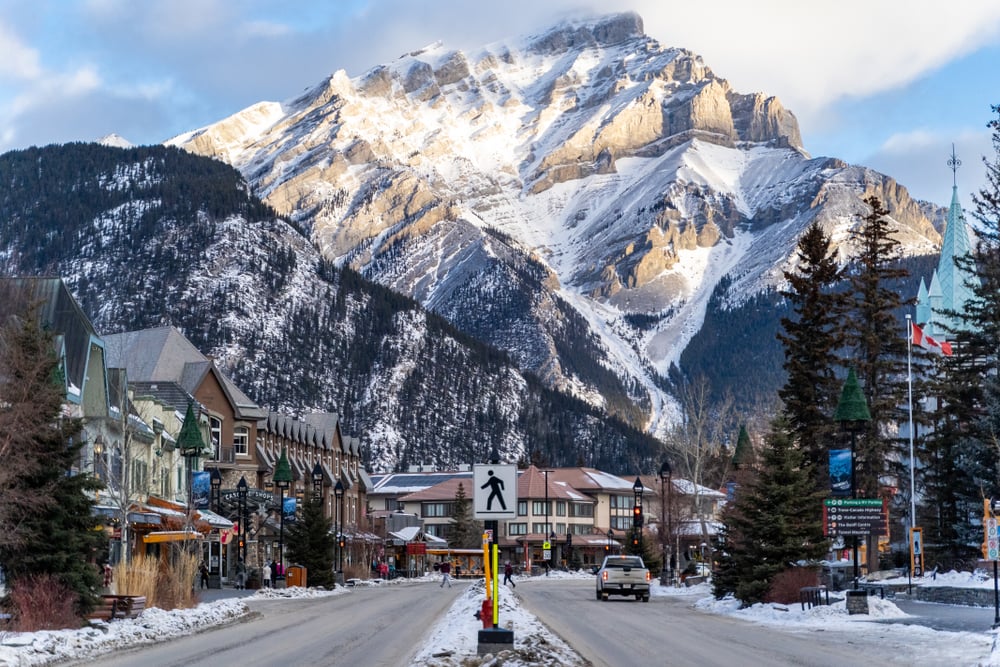 Snowy mountain view over a town street with buildings and trees in foreground, under a partly cloudy sky.