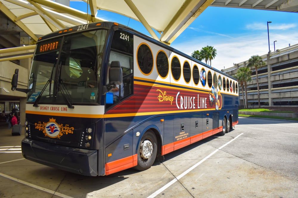 Disney Cruise Line bus parked at airport terminal, with palm trees and parking garage in the background.