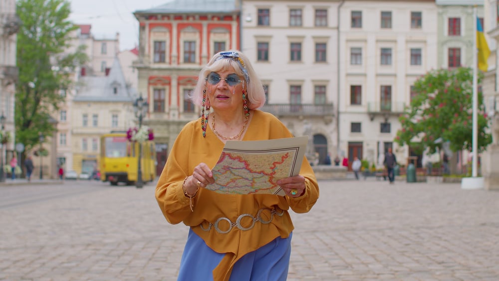 Elderly woman holding a map in a city square with historic buildings and a tram in background.