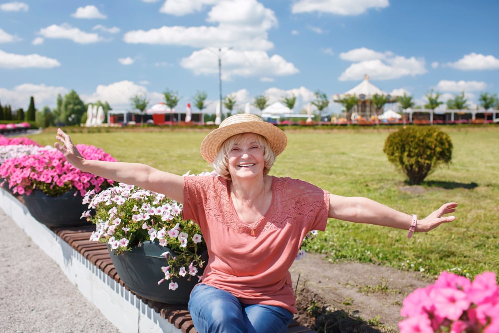Smiling woman with arms outstretched, wearing a hat, sitting on ledge near pink flowers in a park.