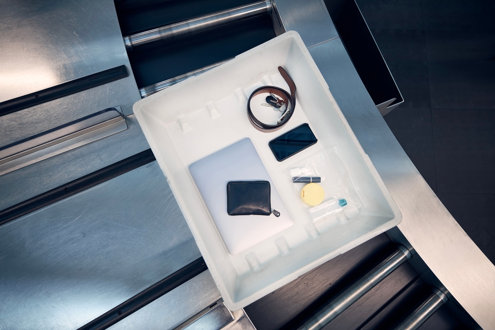 White airport security tray with laptop, phone, wallet, belt, keys, and toiletries bag on conveyor belt.