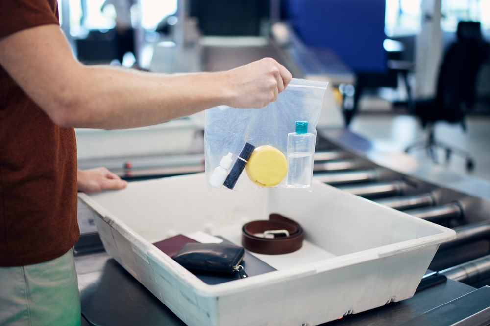 A person holds a clear plastic bag with toiletries over a security tray containing a wallet, belt, and passport.
