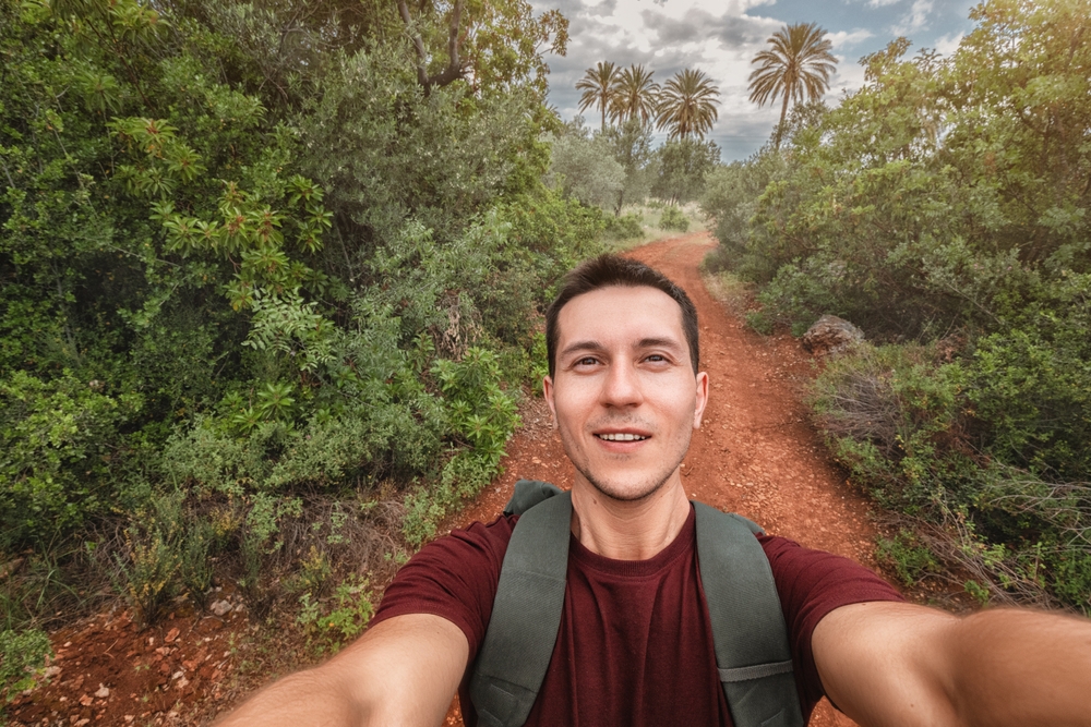 Man taking a selfie on a dirt path surrounded by trees and palm trees in the background.