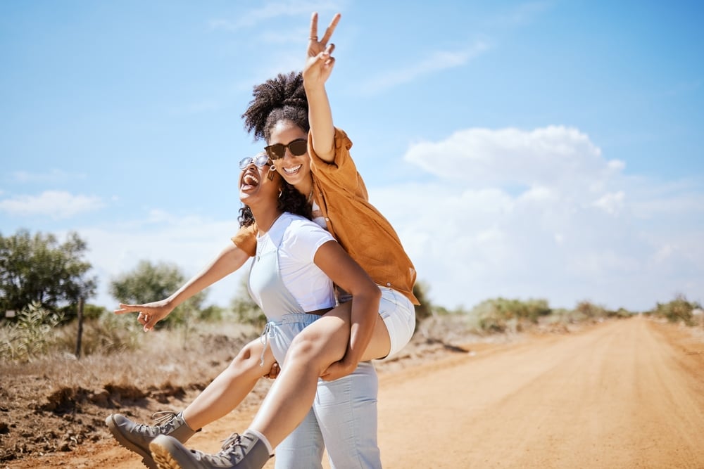 Two people outdoors, one giving the other a piggyback ride, both smiling, with a dirt road background.