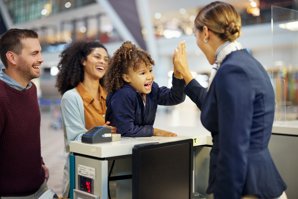 Child high-fives airport staff member at check-in desk, with family nearby smiling.