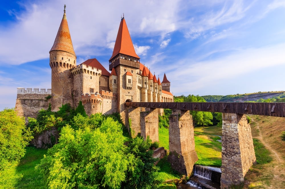 Stone castle with red towers overlooking a wooden bridge, surrounded by green trees under a blue sky.