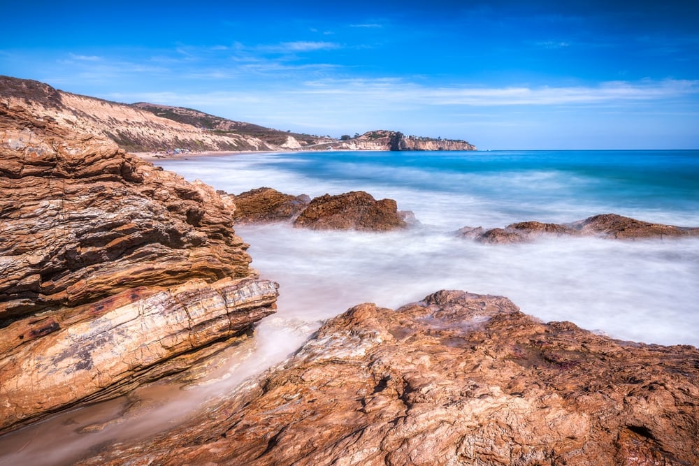Rocky shoreline with waves crashing, blue ocean, and distant cliffs under a clear blue sky.