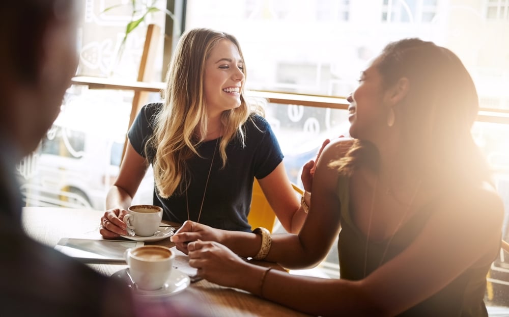 Two women sitting at a café table, smiling and holding cups of coffee.