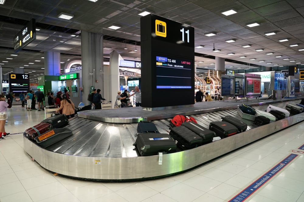Luggage on airport baggage carousel 11, with passengers and digital flight information display in the background.