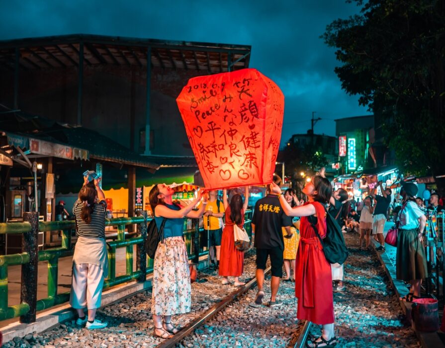 Several people release a red lantern with inscriptions on a railroad track during a nighttime festival.