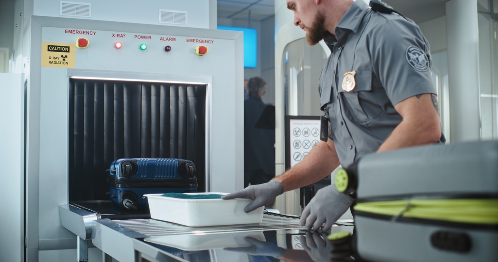 Security officer stands by luggage scanner with suitcase on conveyor belt.