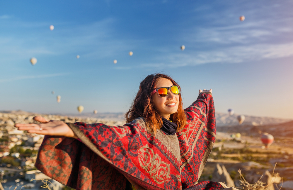 A woman wearing sunglasses and a poncho stands outdoors with arms outstretched, hot air balloons in background.