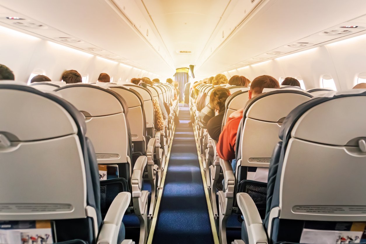 Interior of an airplane cabin with rows of passengers seated, facing forward.