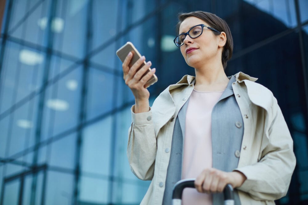A woman in glasses holds a smartphone and suitcase, standing in front of a glass building.