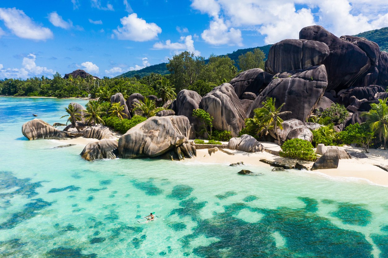 Clear turquoise water, large granite boulders, palm trees on sandy beach, and a person kayaking near shoreline.