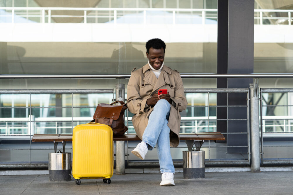 Person sitting on a bench with a yellow suitcase, brown bag, using a smartphone, in an airport.