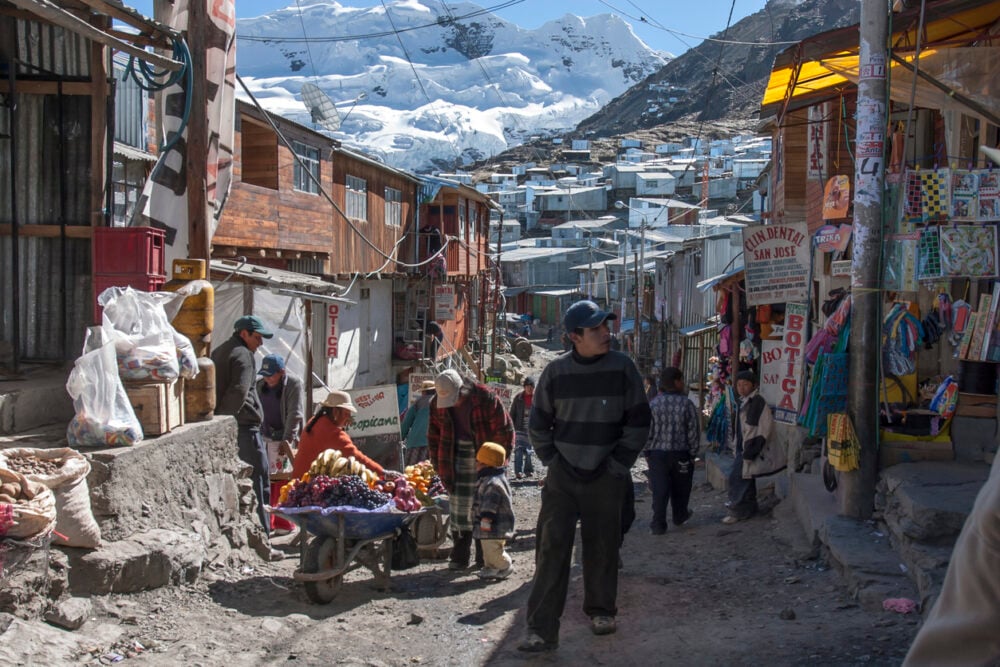 Narrow street with people, market stalls, colorful produce, and snow-capped mountains in the background.