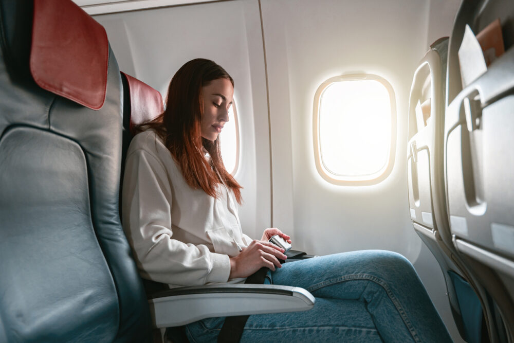 Woman sits alone in airplane seat, looking at phone, beside a bright window.