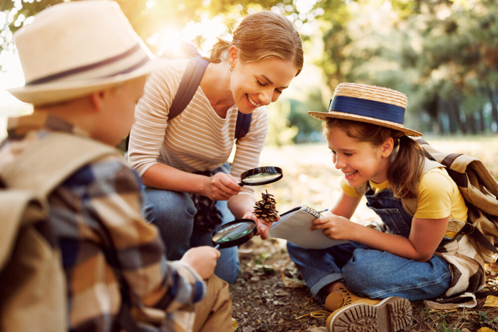 Woman and two kids examining a pine cone with magnifying glasses outdoors.