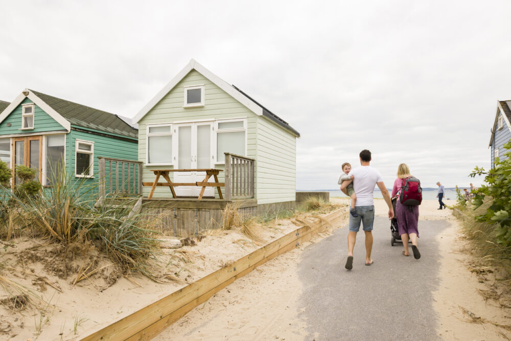 Family walking on a sandy path by beach huts, with a child in arms, towards the sea.