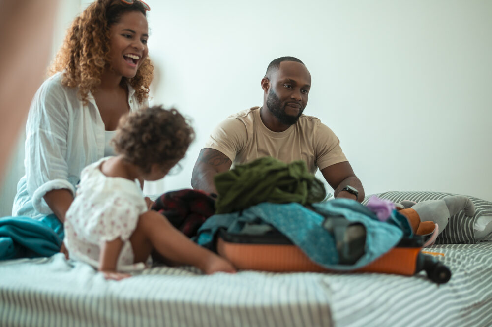 Family sitting on bed, smiling, with suitcase open and clothes scattered around.