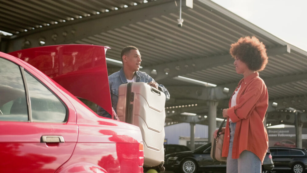Man lifting suitcase from car trunk, woman standing nearby under covered parking area.