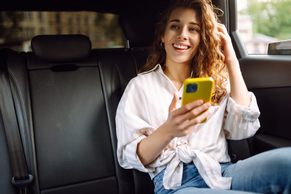 Woman with curly hair holding yellow phone, sitting in car backseat, wearing striped shirt, looking pleasantly surprised.