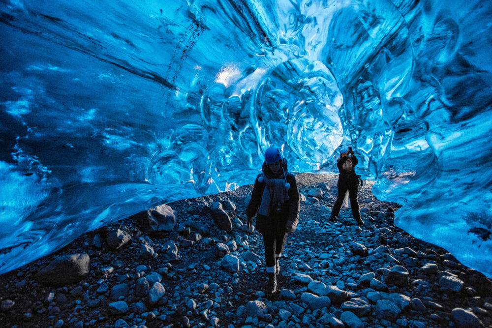 Two people walk inside an illuminated blue ice cave with rocky ground.