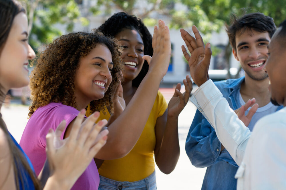 A group of five people happily high-fiving outdoors, surrounded by trees.