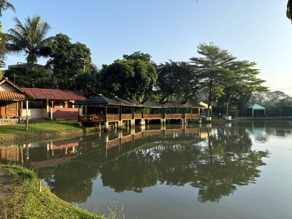 Lakeside pavilion with red roofs and lush greenery, reflecting in the calm water under a clear blue sky.
