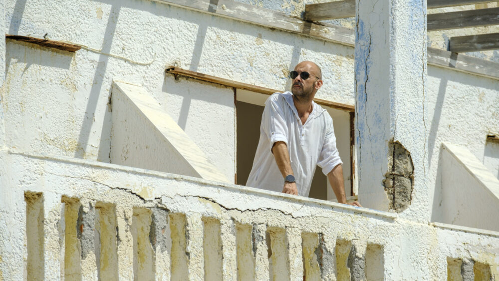 A man in sunglasses leans on a white, weathered balcony railing, looking into the distance.