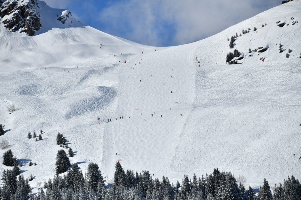 Skiers descending a snowy mountain slope with trees and scattered rocks under a partly cloudy sky.