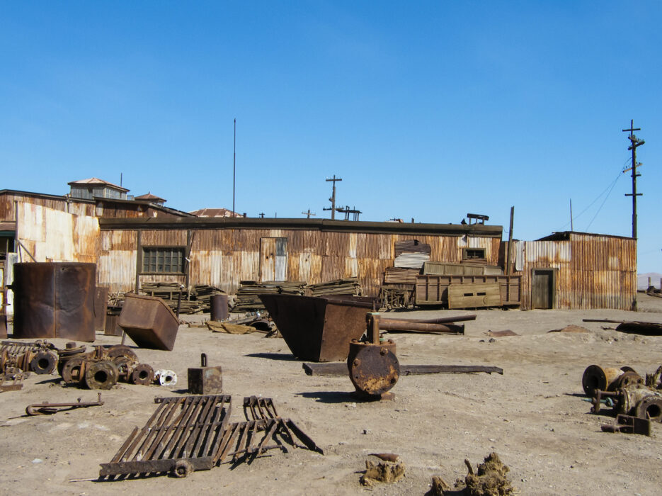 Rusty industrial equipment and scrap metal scattered in front of an old corrugated metal building under blue sky.