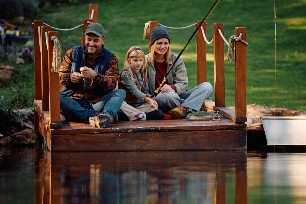 Family sitting on a wooden dock, fishing by a pond, with greenery in the background.