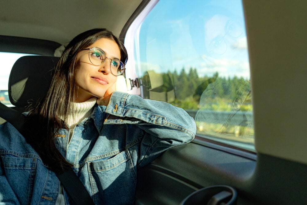 Woman wearing glasses, sitting in car, gazing out window with reflection visible.