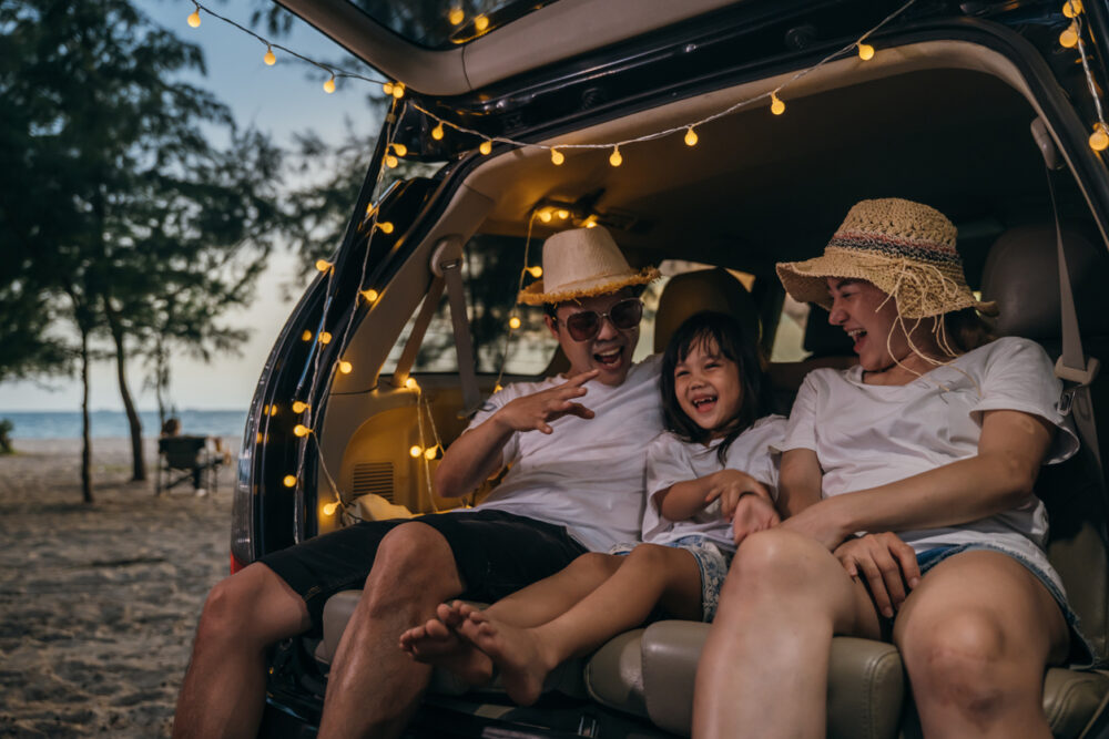 Family sitting in car trunk, decorated with string lights, wearing straw hats, laughing near beach at sunset.