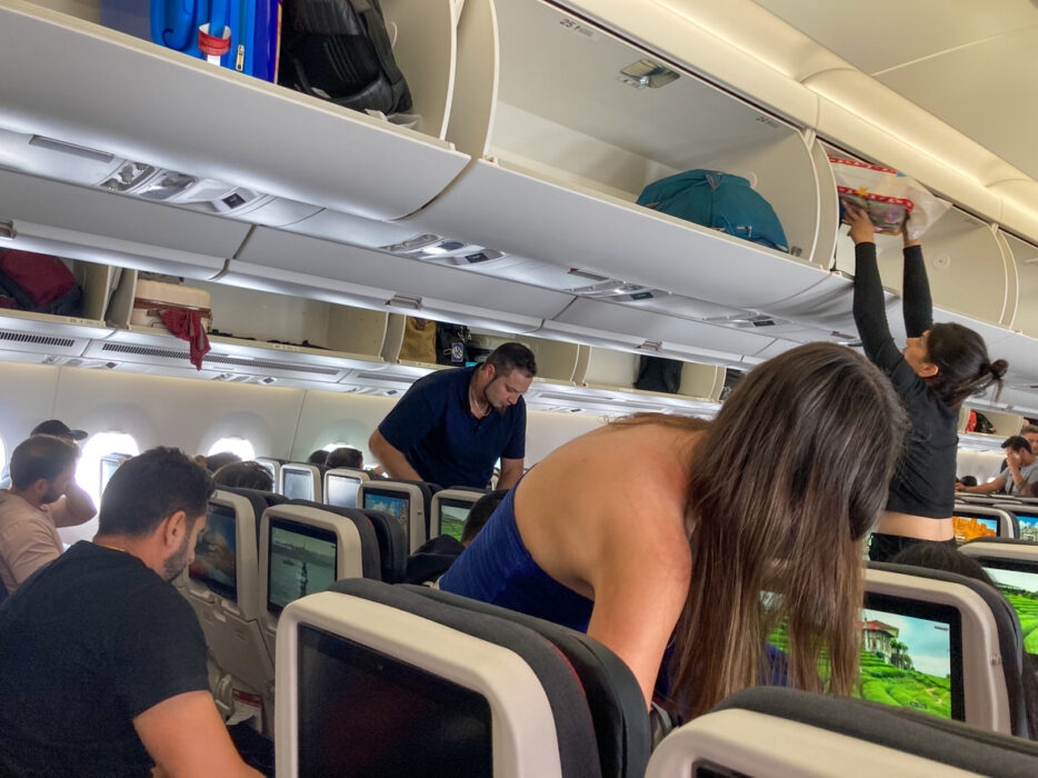 Passengers in an airplane cabin storing luggage in overhead compartments above their seats.
