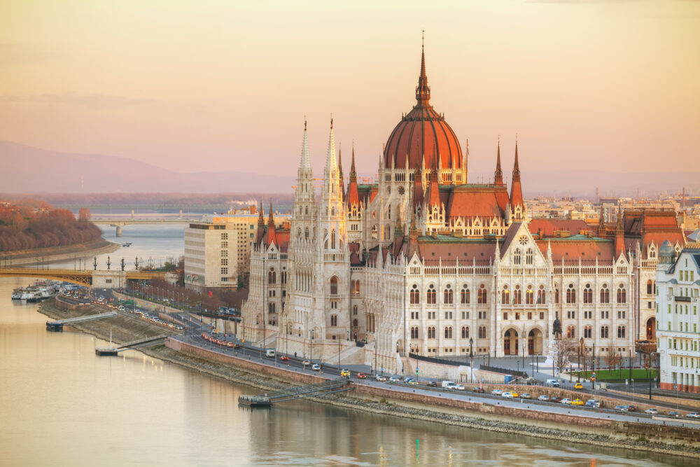 Gothic-style building with multiple spires and domes near a river, set against a sunset sky.