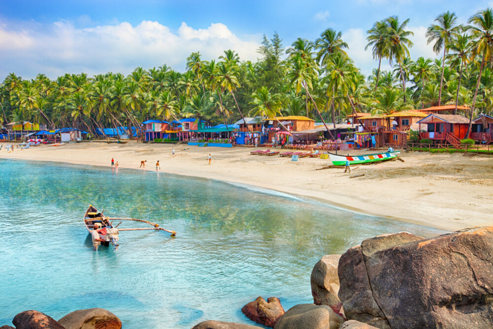 Colorful beach huts, palm trees, boats, and people on a sandy beach with calm blue water.