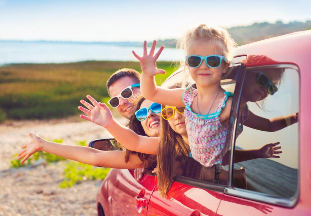 Family wearing sunglasses smiling and waving from car windows by the beach.