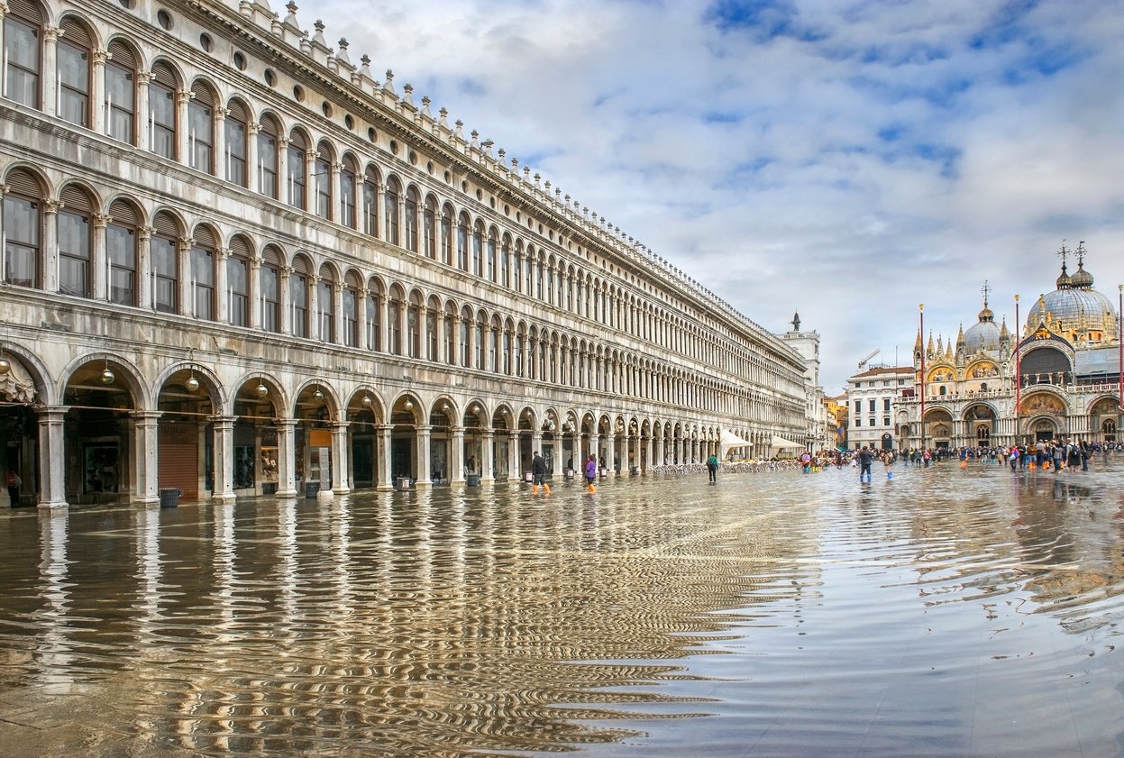 Flooded St. Mark's Square in Venice, with arched building and cathedral in the background.