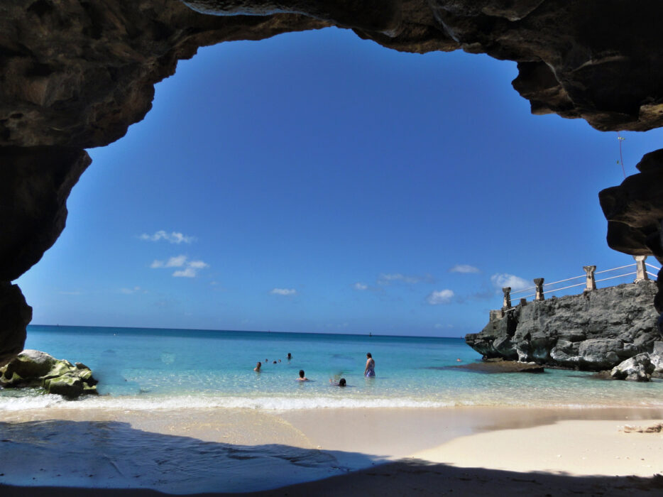 View from cave showing beach, clear water, and people swimming under a bright blue sky.