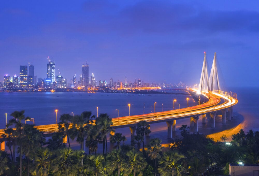 Illuminated bridge extending over water towards city skyline at dusk, surrounded by palm trees.
