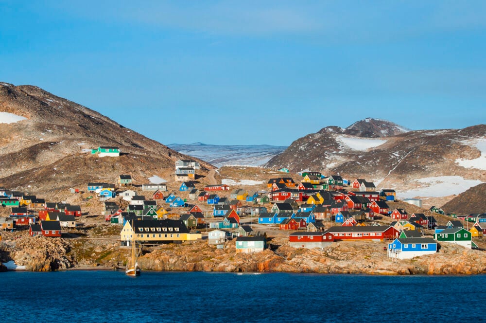 Colorful houses on rocky hillside by blue water, with snowy mountains in the background under clear sky.