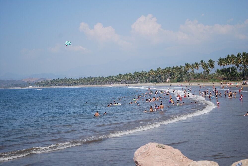 People enjoying the ocean and beach, with parasail and palm trees in the background under a blue sky.