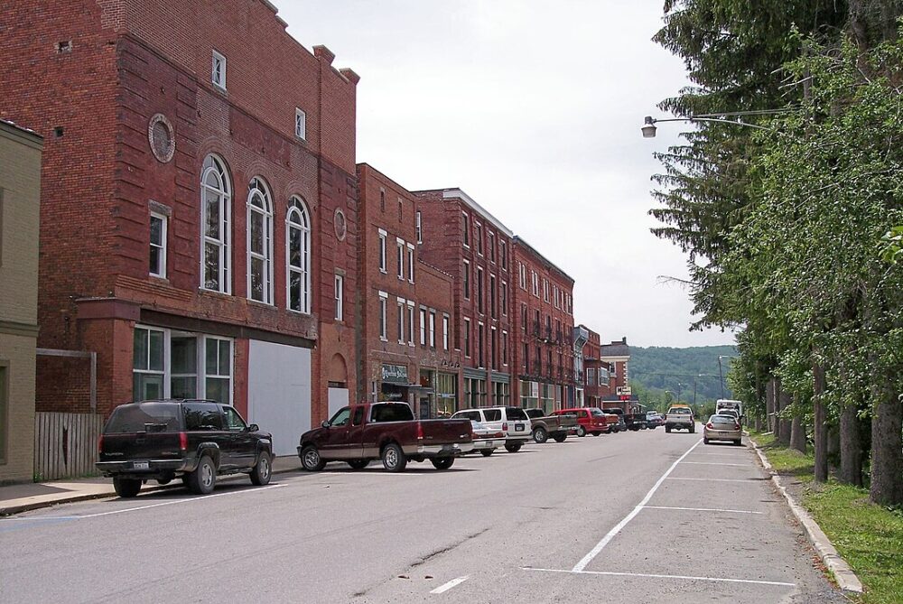 Street with parked cars and red brick buildings on left, trees and lampposts on right, cloudy sky above.