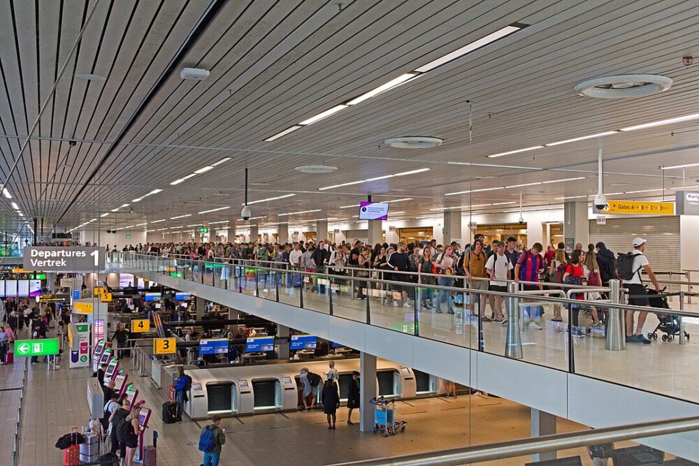 Crowded airport terminal with passengers in line on an upper level, check-in kiosks below, signs displaying directions.